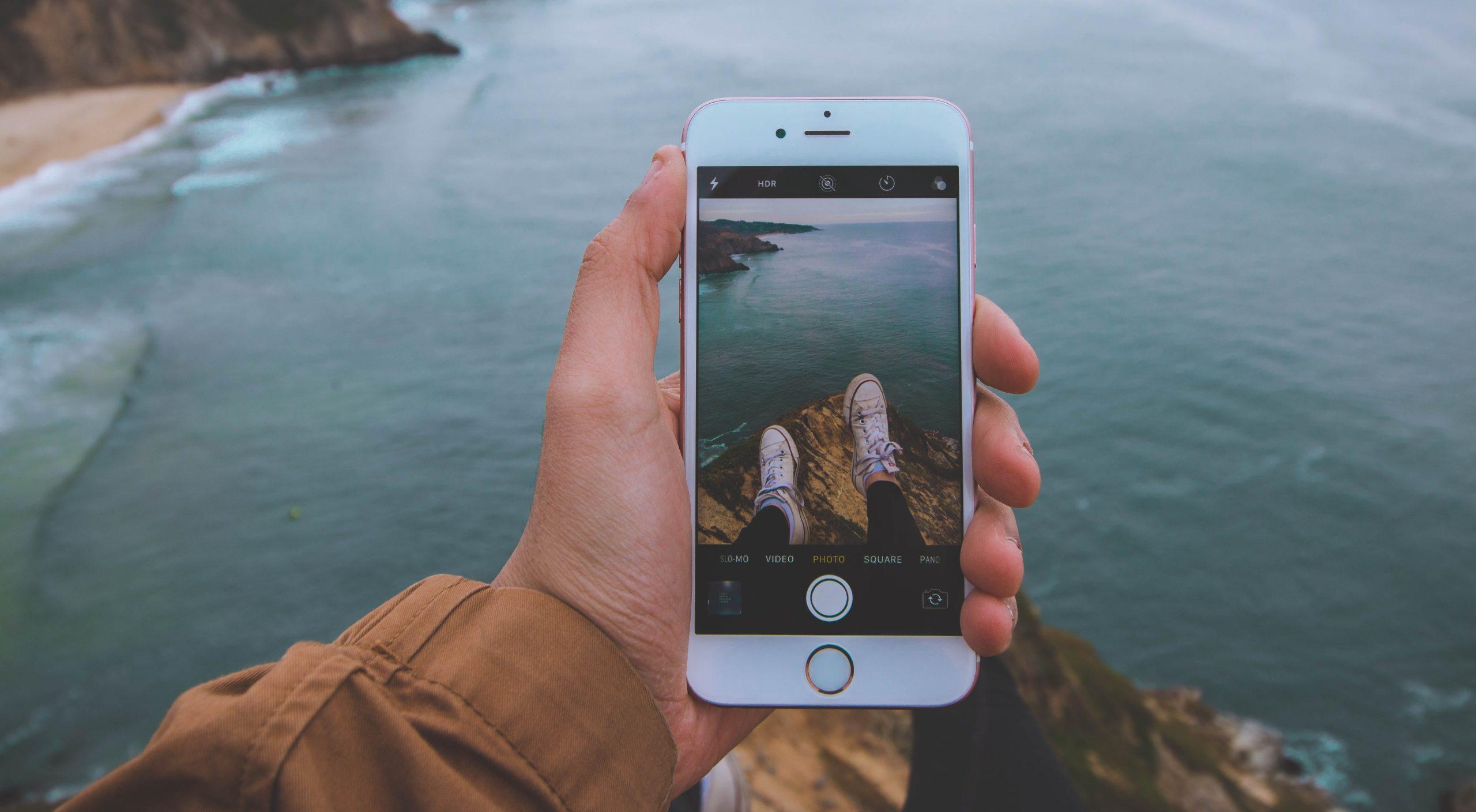 Person taking a photo on an iPhone while sitting on a cliff-face overlooking water. 