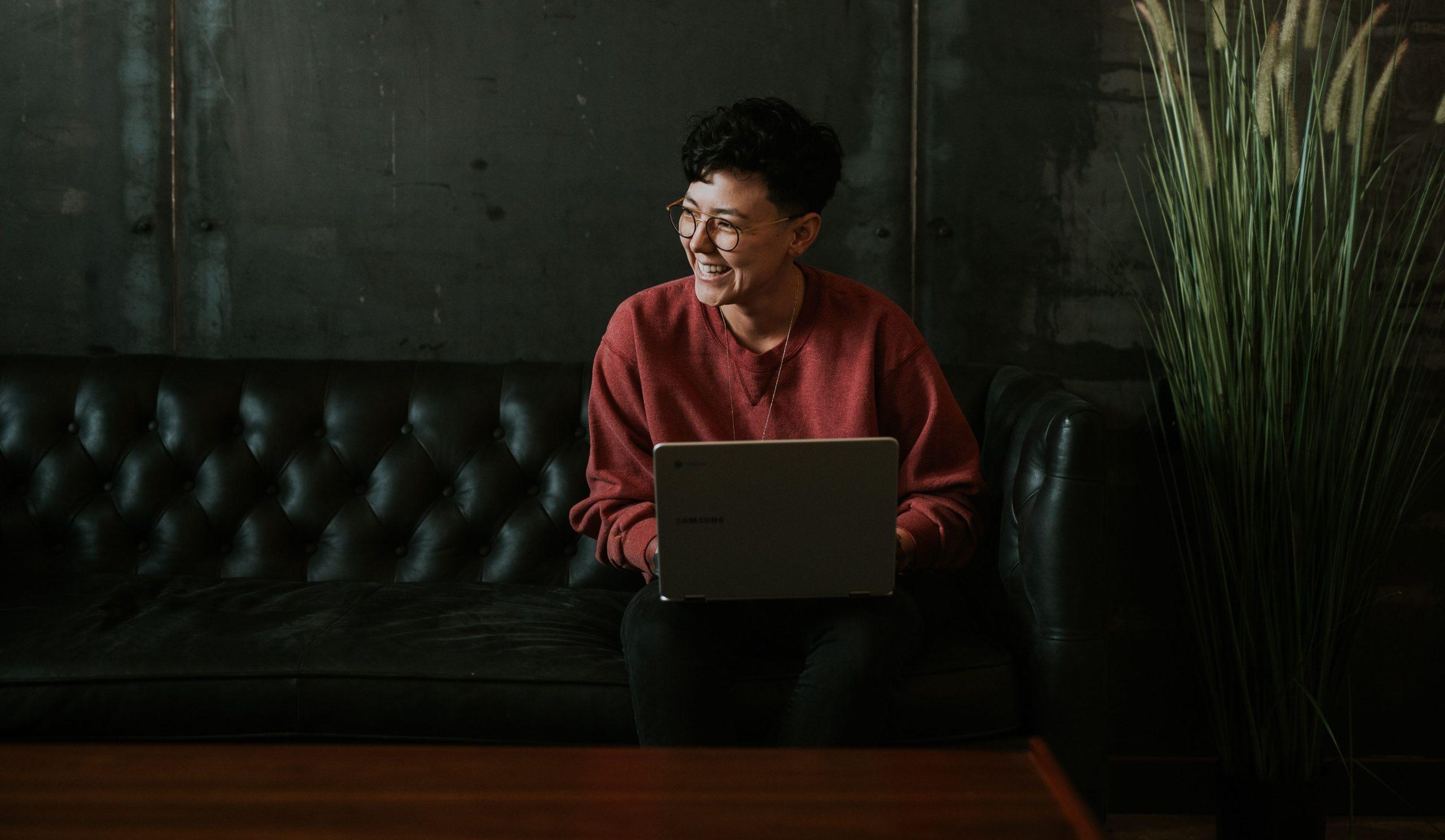 Woman smiling while sitting on couch with laptop in front of her.