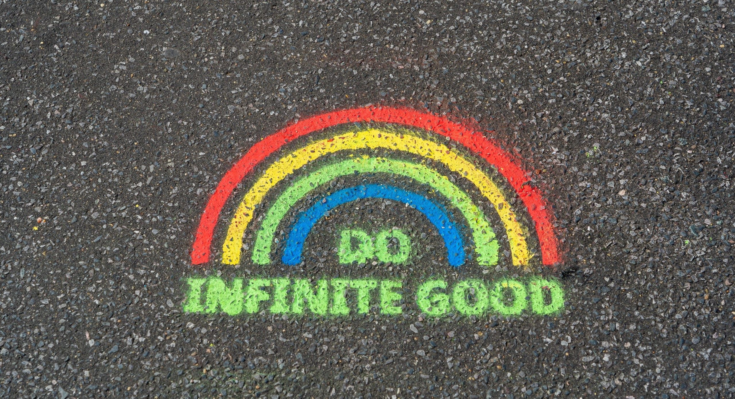 A rainbow drawn in chalk on an asphalt surface accompanied with the words 'Do infinite good'.