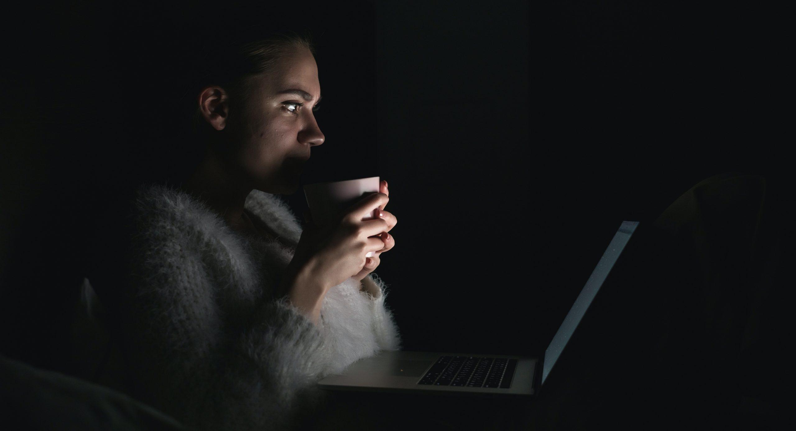 Woman holding a cup while staring at a laptop in a dark setting.