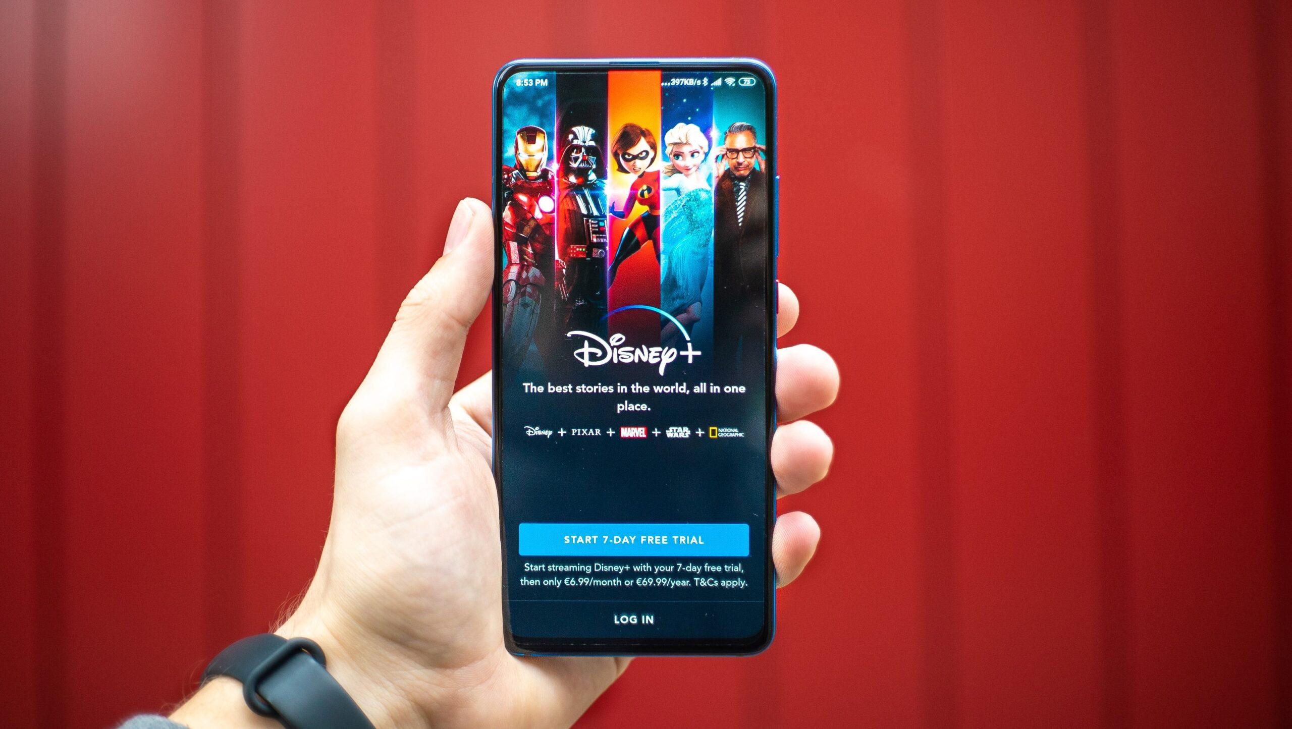 Close up of a hand holding a smartphone displaying the Disney+ app homepage against a red background.