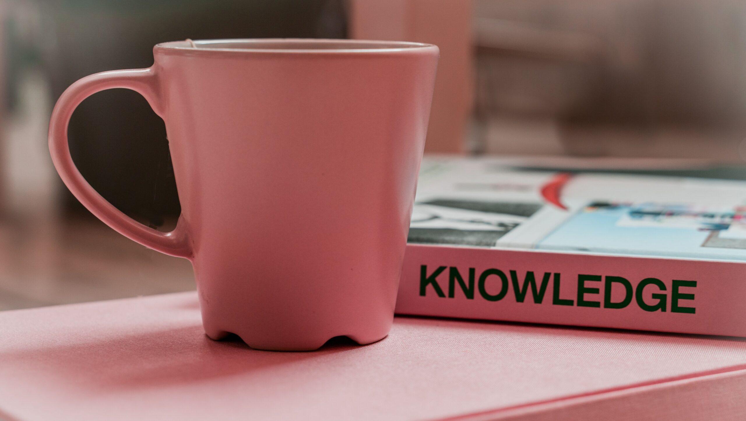 A pink-coloured mug placed on a book in front of another book that has the word 'knowledge' written in its spine.