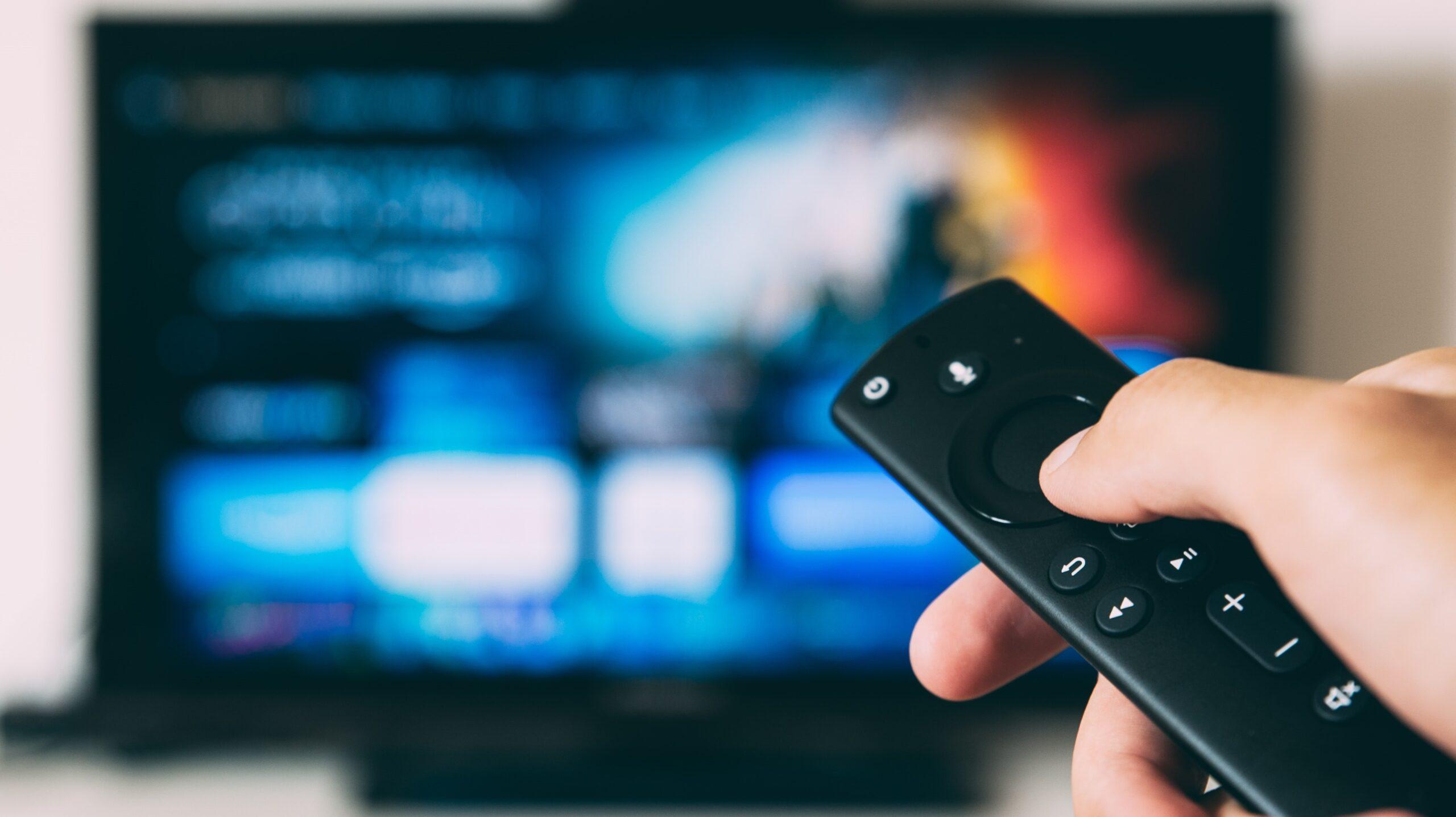 Close up of someone's hand holding a television remote control with a blurred television in the background.