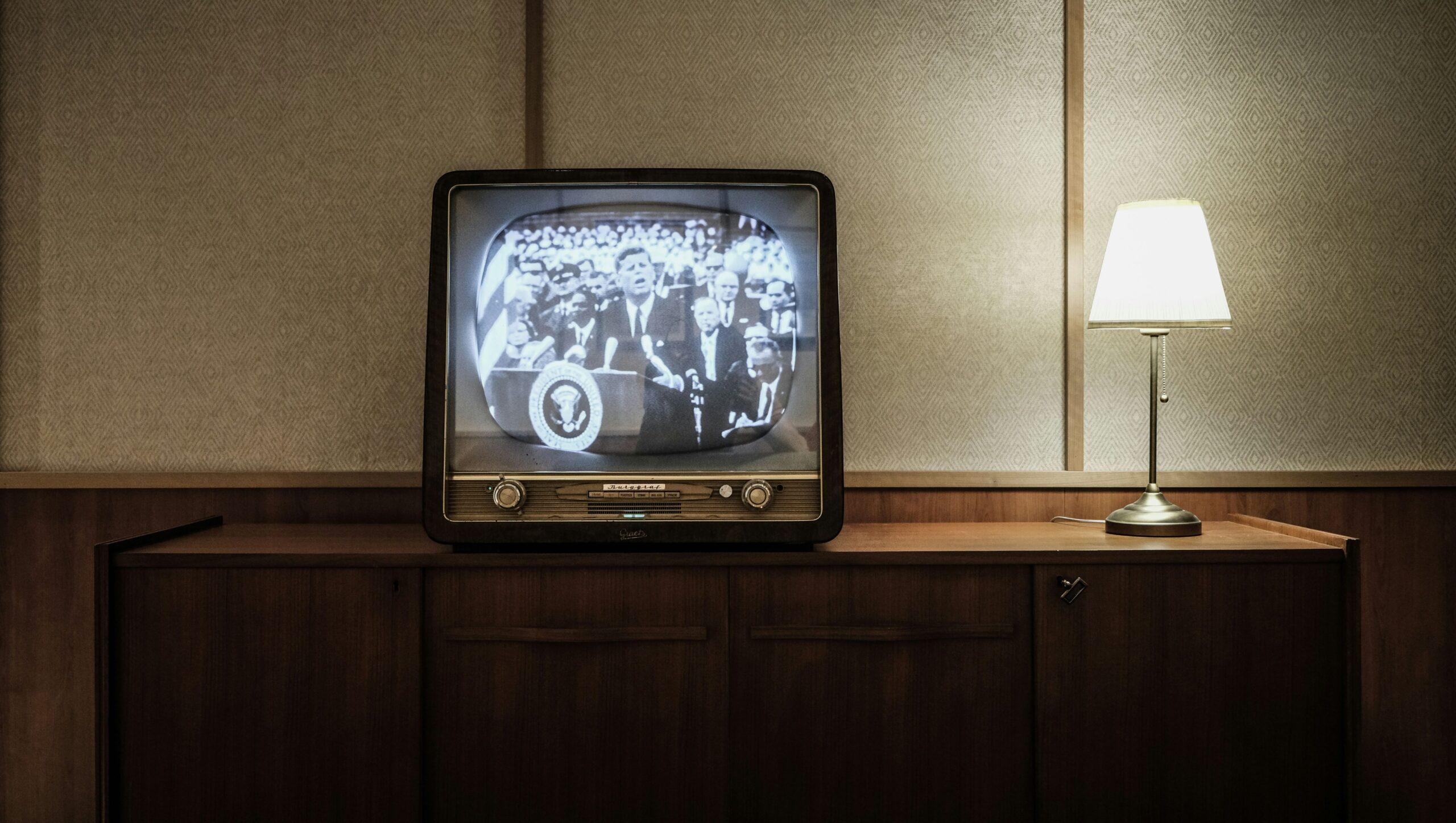 An old black and white television perched on a cabinet next to a lamp. 