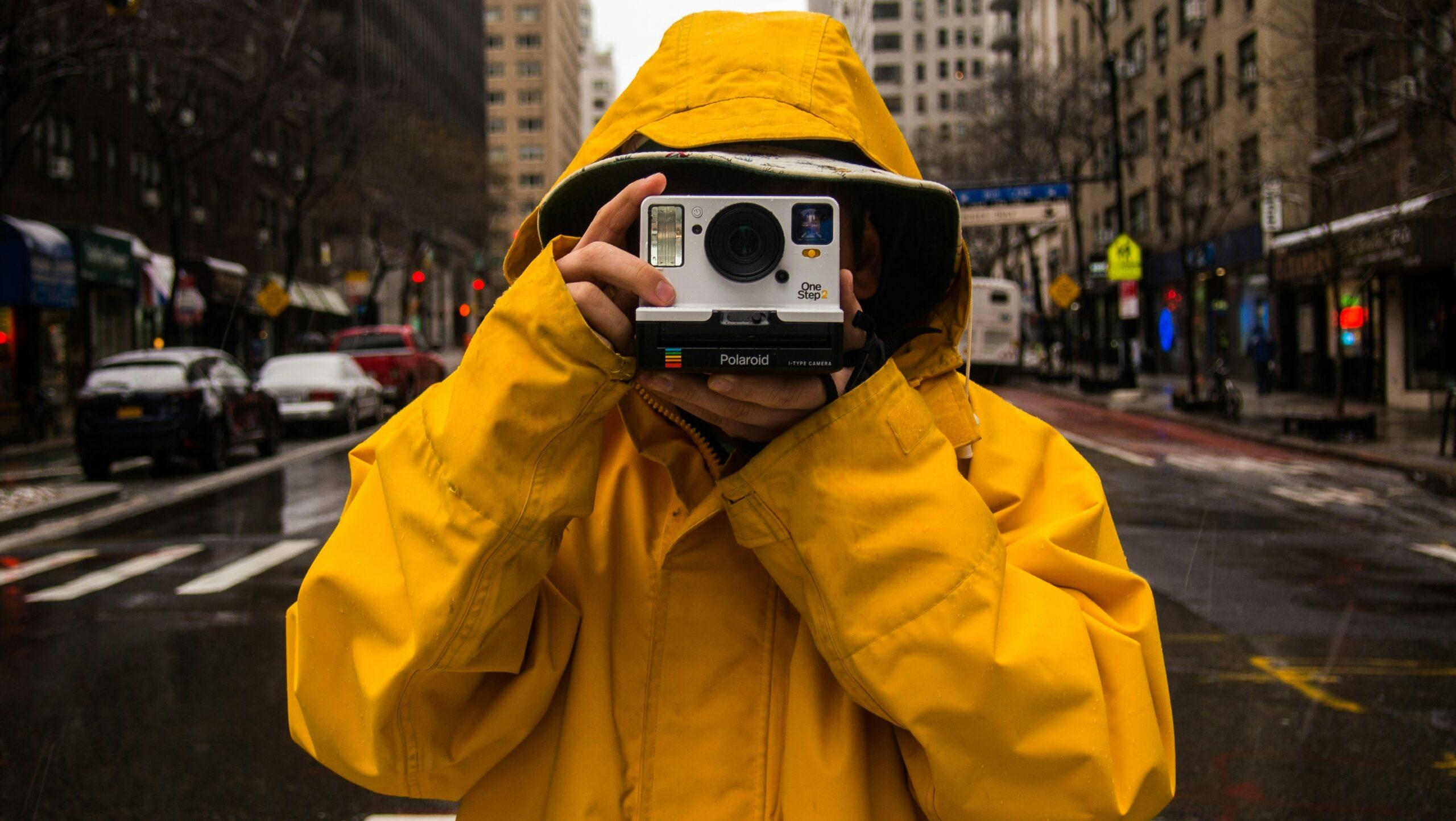 Person dressed in a yellow raincoat standing in the middle of a road while holding a Polaroid camera.