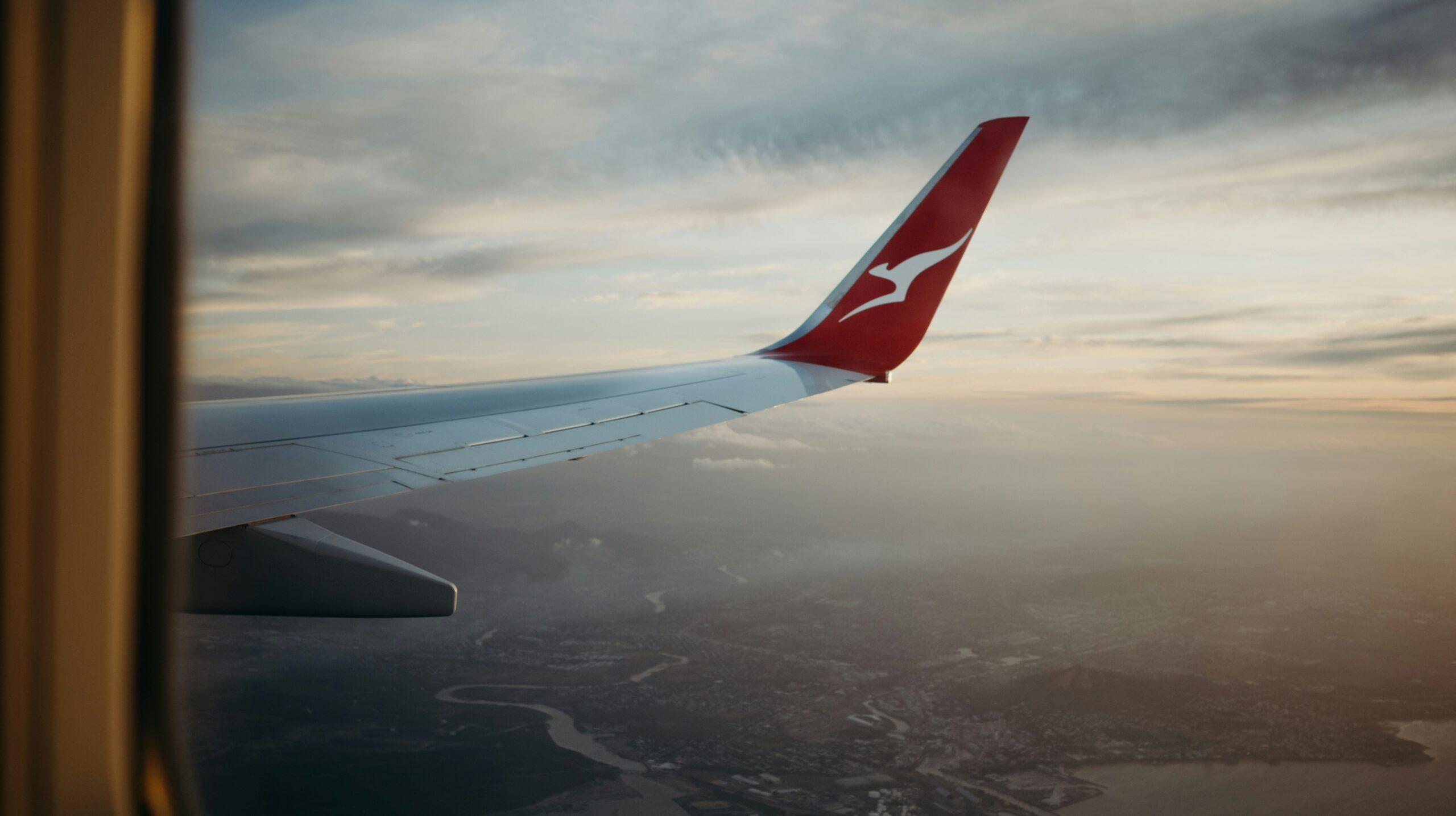 The wing of an air bound Qantas aeroplane backdropped by a colourful sky and overlooking an unidentified location. 